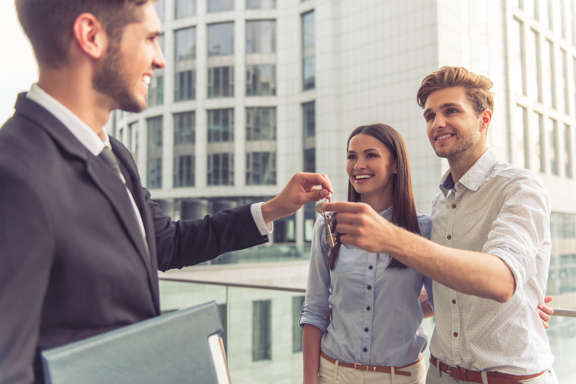 Handsome young businessman is giving keys to happy smiling young couple, modern buildings in the background
